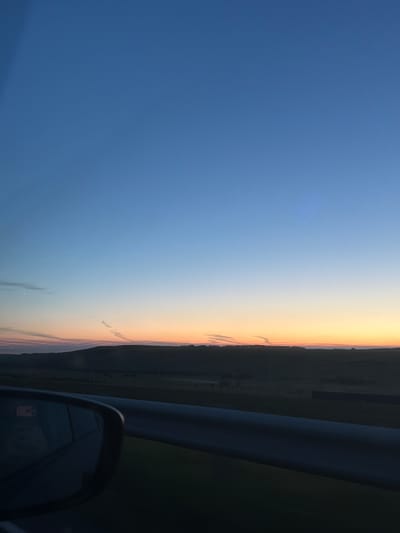 The image captures a serene landscape at dusk, likely in the vicinity of Шуваево, Russia. The sky transitions from a deep, clear blue at the top to hues of orange and yellow near the horizon, suggesting the sun has recently set. Faint contrails from an aircraft are visible, stretching across the twilight sky. Below the sky, rolling hills and a dark, silhouetted treeline form the middle ground. In the foreground, a car's side mirror is visible on the left, reflecting a dimly lit interior. A metallic railing runs horizontally across the lower portion of the frame, indicating the perspective is from inside a moving vehicle. The overall impression is one of peaceful travel during a transitional time of day.