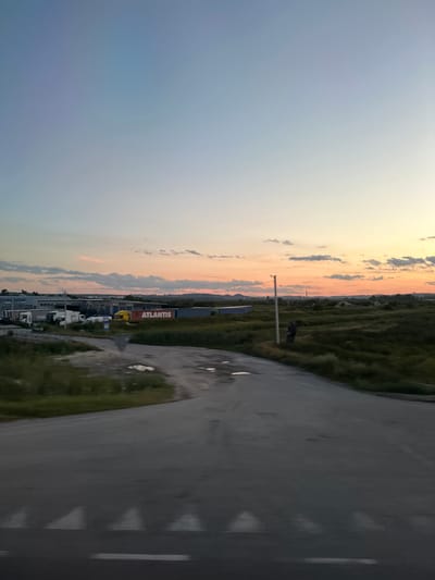 The image depicts a twilight scene in Kamianopil, Ukraine, featuring a paved road in the foreground leading towards a field with scattered buildings and vegetation. The sky exhibits a gradient of blue to orange hues, indicating sunset. In the mid-ground, several industrial-style buildings are visible, with a prominent sign reading "ATLANTIS". A yellow truck is parked near this building, alongside other white trucks and trailers. A solitary utility pole stands in the middle of a grassy area to the right of the road. The terrain is uneven, with some patches of wetness on the road surface. A blurred white line, possibly a pedestrian crossing, is visible at the bottom of the frame. The overall impression is that of an industrial or commercial area on the outskirts of the city during dusk.
