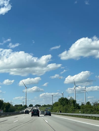 The image depicts a view from a vehicle traveling on a highway near Würges, Germany. The sky is blue with scattered cumulus clouds. Several wind turbines stand near the roadside. On the right, multiple vehicles are traveling in the same direction on the highway. Trees line the edge of the road.
