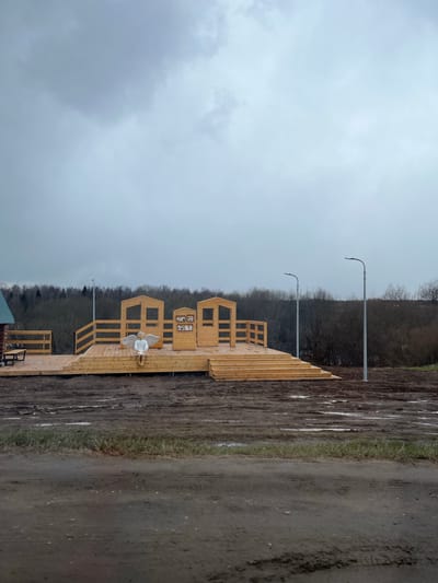 The image depicts an outdoor scene in Чутырь, Russia, likely during late autumn or early spring, characterized by bare trees and muddy terrain under an overcast sky. The sky is uniformly grey, suggesting a dull, possibly cold day.
The primary subject is a newly constructed or well-maintained light-colored wooden deck or walkway that extends across the midground. This wooden structure features railings and several small, house-like facades built upon it. Some of these facades have open, rectangular window-like frames. One facade in the center displays a series of small framed images, possibly of cartoon animals or decorative figures, within its opening. A white statue or figurine of an angel, complete with large wings, is prominently seated on the wooden deck, adding a whimsical or contemplative element to the scene.
Beyond the wooden platform, the ground consists of dark, wet earth and mud, marked by distinct tire tracks. Scattered patches of sparse green grass are also visible. In the background, a dense line of bare deciduous trees or bushes forms the horizon line. Two tall, slender street light poles with curved tops extend upwards from behind the tree line, indicating existing or developing infrastructure.
No people are visible, and no specific activity is taking place, creating a quiet and still atmosphere. The overall impression is one of a developing or recently completed recreational area in a rural or semi-rural setting. No legible text is discernible on any objects within the image.