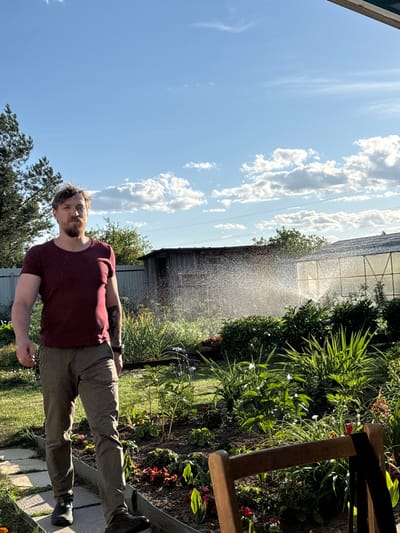 The image depicts a young man standing in a garden under a bright, sunny sky with scattered clouds. He is wearing a maroon t-shirt and khaki pants. His left arm is bent at the elbow and his left hand is resting on his hip. His right arm is extended slightly, with his hand open. He has a beard and short, light brown hair. A tattoo is visible on his left arm.
The garden setting features lush green foliage, some flowering plants with red and white blooms, and dark soil. In the background, a greenhouse structure is visible, and water is being sprayed, creating a fine mist that catches the sunlight. A wooden bench is partially visible on the right side of the frame. The scene appears to be set during the daytime, with the sun casting shadows and illuminating the water spray. The overall mood seems relaxed and summery. No visible text is present in the image. The location context is Sarov, Russia.