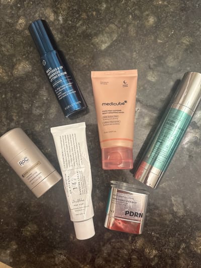 The image shows a collection of skincare products on a dark, possibly marbled countertop. The products include: VT PDRN Essence Stick Balm, Medicube PDRN Pink Caffeine Night Wrapping Mask, CICA x HYALON PDRN Reedle Shot, Allie’s of Skin Multi Peptides & GF Advanced Lifting Serum, Dr. Althea 147 Barrier Cream, and RoC Derm Correxion Firming Serum Stick. The labels indicate ingredients such as PDRN, caffeine, peptides, retinol, hyaluronic acid, and ceramides. There are no people visible in the scene. The location in Clifton, United States of America, is not evident from the image.