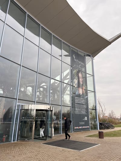 The image displays the exterior of the Therme București building in Săftica, Romania, under an overcast sky. The structure features a modern glass facade with multiple vertical and horizontal panes, topped by a curved, light-colored overhanging roof. Reflections of interior palm trees and the grey sky are visible on the glass. A large, dark vertical banner is affixed to the facade's right side, advertising the "HERBARIUM FESTIVAL" from "4 - 17 MAY 2026," with a graphic of a woman and text detailing "Europe's largest wellness festival."

At the ground level, a silver-framed revolving glass door serves as an entrance on the left. A person wearing a dark jacket and white pants is partially visible, entering this door. To the right of the door, a dark grey rectangular doormat is situated on light brown paved ground. A second individual, male, wearing a dark jacket, dark pants, and shoes with red soles, is walking across this doormat from left to right, looking down at a handheld device. Further to the right, a dark brown cylindrical waste bin stands next to a row of parked bicycles on a patch of grass. Distant bare trees and minimal structures are visible in the background on the right.