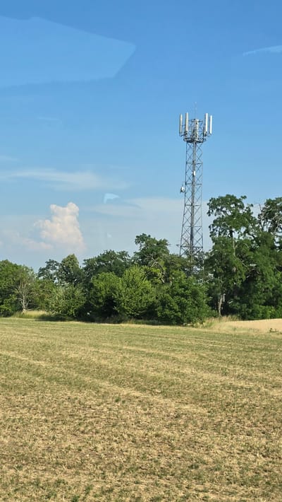 A cell tower stands tall in a rural landscape, set against a bright blue sky with scattered clouds. The tower, a lattice of metal, is topped with multiple antennas. In the foreground, a field of dry, yellowish-brown grass stretches out. Behind the field, a dense line of green trees forms a backdrop, partially obscuring the base of the cell tower. A single fluffy white cloud, shaped somewhat like a figure, is visible in the sky to the left of the tower. The overall impression is one of a clear, sunny day in a predominantly natural setting. This scene is located in the vicinity of Drysice, Czechia.