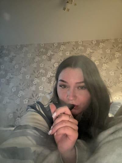 The image shows a close-up, indoor, low-angle selfie of a young woman with dark hair. She is looking towards the camera with a neutral expression, and her mouth is partially open as she holds a dark-colored vape pen to her lips. Her right hand, adorned with a ring and manicured nails, is positioned near her mouth. She is wearing a striped top. The background consists of floral patterned wallpaper and a light-colored ceiling with a fixture visible at the top. The scene appears to be a bedroom or living area. No specific location in Osentsy, Russia can be determined from the image.