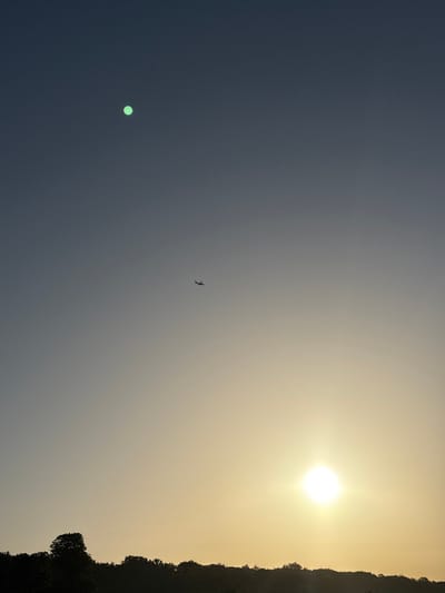 This is a photograph of a sky with a gradient of colors, transitioning from a dark blue on the left to a bright yellow on the right. The sun is a prominent, glowing orb in the upper right quadrant of the image. A silhouetted bird is captured in mid-flight in the upper central area of the frame. In the lower left quadrant, there is a small, circular, light green object that could be lens flare or another celestial body. The right edge of the image is bordered by a dark, silhouetted treeline, suggesting a landscape setting, possibly a forest or hills. The overall lighting and color palette indicate that the photograph was taken during sunrise or sunset. The absence of any people or specific landmarks makes it difficult to pinpoint the exact location within Kongens Lyngby, Denmark, but the natural elements suggest an outdoor scene. The image evokes a sense of natural beauty and the vastness of the sky. There is no visible text in the image.