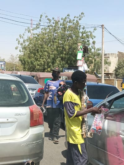 This image captures a street scene in Hotoro, Nigeria, under clear daytime conditions. In the foreground, the rear of a silver Toyota Corolla hatchback is visible, featuring a taillight with reddish-brown tape repair and a scuffed bumper. To the right of the Corolla, a young male, dressed in a yellow t-shirt with dark blue sleeves and dark blue pants, stands alongside a silver car. He holds an upright pole adorned with various small items, likely keychains or trinkets, and extends a clear plastic bag containing numerous colorful, small cylindrical objects, possibly lip gloss or candy, towards the car's passenger compartment. Behind this individual, a man wearing a blue camouflage-patterned t-shirt, dark pants, a dark baseball cap, and a black face mask walks towards the right. He carries a dark bag with "REEBOK" branding and a white object in his hands.
The midground and background feature multiple vehicles, including several light brown three-wheeled auto-rickshaws (keke napep) and sedans, suggesting traffic. A prominent green tree with dense foliage occupies the central background. Overhead power lines and a utility pole with attached wiring and various mounted items are visible. A partially obscured billboard in the left background displays text, including "THE NIGERIA." Two red lattice transmission towers are visible in the distant background, rising above the tree line. The ground appears to be paved.