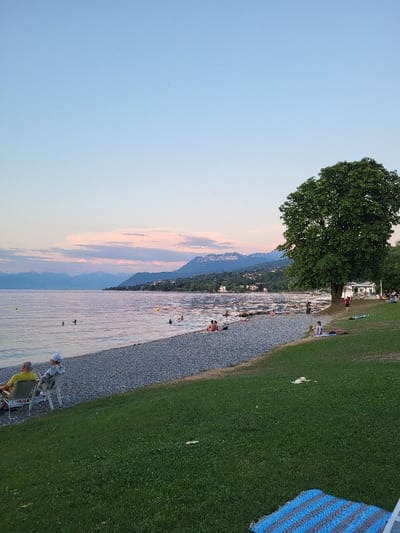 A serene landscape features a calm body of water stretching towards the horizon, bordered by a pebbled beach. People are scattered along the shoreline and swimming in the water. Lush green grass extends to the right of the beach. A large tree stands near the shore. In the distance, mountains rise, and the sky transitions from a clear blue to a soft, pinkish hue, possibly during sunset.