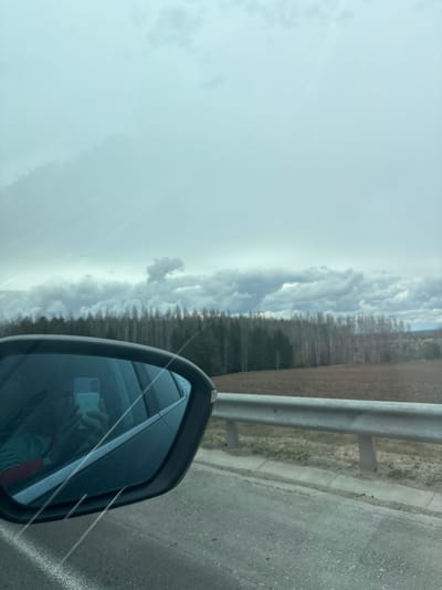 The image, correctly oriented, depicts a view from the passenger side of a vehicle, looking out across a rural landscape under an overcast sky. In the immediate foreground, the black frame of a car's side mirror is prominent, reflecting a person holding a light blue/green smartphone with a dual-camera system, likely taking the photo. The person appears to be wearing a light-colored long-sleeved top with a subtle red or orange garment underneath.

Outside the vehicle, to the right, a gray asphalt road or track runs alongside a sturdy metal guardrail. Beyond the guardrail, a strip of sparse, dry vegetation gives way to an expansive field of light brown earth or dry grass. Stretching across the midground is a dense, dark line of tall, slender trees, appearing somewhat silhouetted against the bright sky, suggesting a forest or treeline on the horizon. The trees appear to be largely bare, indicating late autumn, winter, or early spring.

The sky, occupying the left and upper portions of the image, is filled with dramatic, heavy gray and white clouds, signifying an overcast day. Some faint light streaks, possibly sun rays attempting to penetrate the clouds or lens flares, are visible in the upper left. The overall scene suggests a journey through a rural area, consistent with the given location context of Khutor, Russia, with the weather appearing cool and cloudy. No discernible text is visible apart from the general design of the smartphone.
