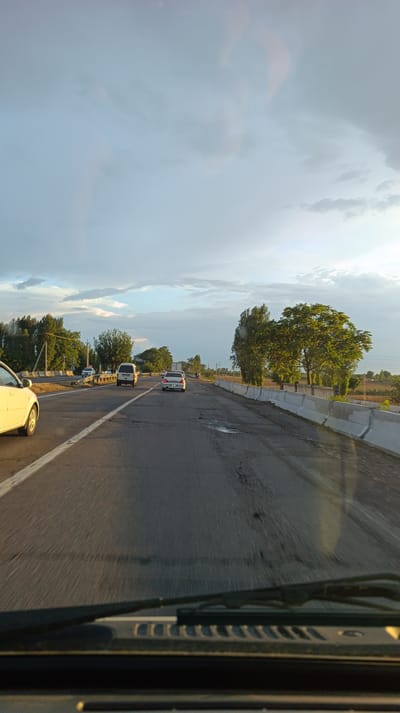 The image shows a view from inside a vehicle on a road in Iyazbagish, Uzbekistan. The road has multiple lanes and appears to be in fair condition with some potholes visible. Concrete barriers line the right side of the road. Several vehicles are on the road, including a white car in the left lane, a white van in the center lane, and a white sedan in the right lane. Trees and vegetation are visible on both sides of the road, with some buildings and fields in the background. The sky is partly cloudy with a mix of blue sky and grey clouds, suggesting it might be late afternoon or early evening. The sun is casting a warm light on the scene. The view is from the perspective of the driver, with the windshield and wipers visible in the foreground.