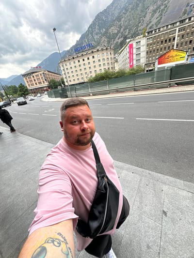 The image, originally rotated 90 degrees counter-clockwise, depicts a man taking a selfie on a city street in Andorra la Vella, Andorra. The man, centrally framed, wears a light pink t-shirt and a black fanny pack or crossbody bag across his chest. He has a beard and short dark hair, looking directly at the camera with a neutral expression. A tattoo visible on his left forearm reads "enjoy".
The background features a multi-lane asphalt road with white markings, leading into the distance. Alongside the road, various buildings line the street. Notable signs include "NOVOTEL" on a distant building and "Mercure HOTEL" on a building closer to the foreground. Further down the street, other commercial signs are visible, displaying "Recinte Firal de l'Automobil," "Montes Autos," "Tel. 86 19 20 - Fax. 86 19 30 - Mob.," and "COMPRA I VENDA DE VEHICLES." Another sign reads "Bingo."
In the far background, imposing mountains rise under a cloudy, daytime sky, highlighting Andorra's mountainous landscape. The scene indicates an urban environment integrated with natural surroundings. Another person in a dark coat can be seen walking on the sidewalk further down the street.