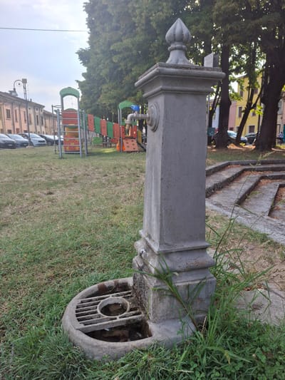 In the city of Pieve Saliceto, Italy, an old stone water fountain stands in a grassy area next to a playground. The fountain, made of weathered grey stone, features a rectangular base with a decorative cap. A metal spigot protrudes from the front of the fountain. The ground around the fountain is covered in green grass, with some patches of dry earth. Behind the fountain, a colorful playground with slides and climbing structures is visible. Several cars are parked in the background, and some residential buildings line the street. Trees with lush green foliage provide shade, and the sky is partly cloudy. The scene appears to be an outdoor public space, likely a park or a town square, capturing a moment of everyday life in Pieve Saliceto.