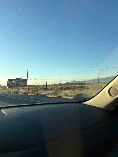 This media file captures a landscape view through a car window, oriented vertically. The scene depicts a desert environment with dry vegetation, mountains in the distance, and a clear blue sky. A billboard is visible alongside a road, featuring advertisements with text. Power lines and utility poles are scattered across the landscape, suggesting a semi-rural or roadside setting. The time of day appears to be late afternoon or early evening, indicated by the warm, low-angled sunlight casting long shadows and illuminating the scene with a golden hue. The presence of the car interior, including part of the dashboard and a speaker grille, confirms the perspective is from inside a vehicle. No people are visible in the frame. The overall mood is one of travel and observation of a vast, open landscape.

The billboard's text is partially obscured but appears to include the word "ROUGH" and possibly "STONES." There are also images of people on the billboard.