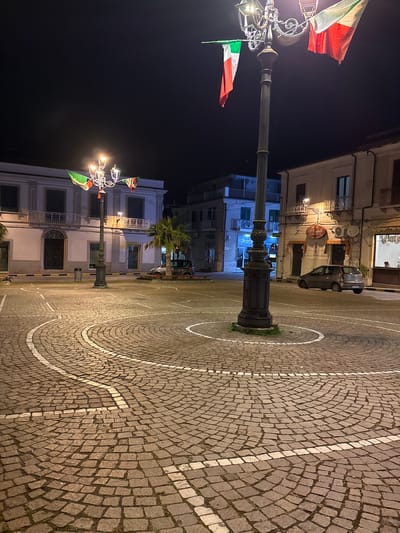 Captured at night, the image reveals a public square in Polistena, Italy. The square is paved with cobblestones in a circular pattern, accentuated by concentric white lines. At the center stands a vintage lamppost adorned with Italian flags. The surrounding buildings are multi-story structures with windows, balconies, and subtle architectural details. One building showcases a storefront with a sign above it. A car is parked near one of the buildings. A palm tree adds a touch of greenery to the scene. The sky is dark, indicative of the nighttime setting. The scene is static, with no visible people or immediate actions taking place.