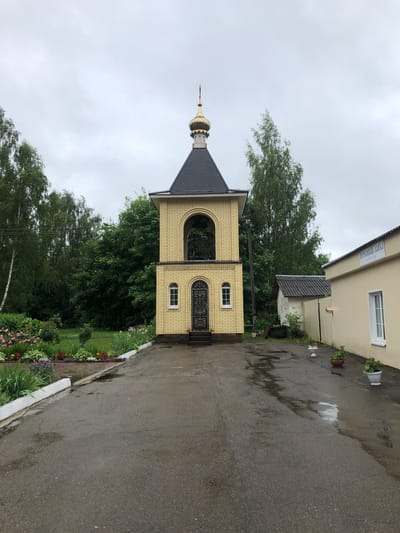The image shows a bell tower in Safonovo, Russia, on a cloudy day. The bell tower is made of yellow brick and has a black roof with a golden dome. In front of the bell tower, there is a paved walkway with some puddles, indicating recent rain. To the left of the walkway, there is a flower bed with various colorful flowers. To the right of the bell tower, there is a long, light-yellow building with a sign that reads "ЦЕРКОВНАЯ ЛАВКА" (Church Shop). Behind the bell tower and the shop, there are several trees and a smaller white building. There are no people visible in the image.