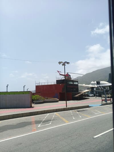 This media file shows an outdoor scene in Macuto, Venezuela, on a bright, sunny day with scattered clouds. The foreground features a paved road with parking markings. A concrete curb separates the road from a pedestrian walkway. Behind the walkway are several large, stacked shipping containers. The containers are red and grey with a green top on one. A small area of greenery is visible between the red and grey containers.
In the middle ground, a multi-level structure built on top of shipping containers serves as a platform for two distinct vehicles. A red and silver helicopter is parked on the upper level of this structure. To the right, the tail and wing of a white airplane are visible, also seemingly integrated into a building or structure. A white car is parked on a ramp leading to the airplane structure. A lamppost with multiple spotlights stands prominently near the helicopter.
In the background, a lush, green mountainside rises dramatically against the sky. The sky is a light blue, indicating daytime.
A sign with blue lettering that appears to say "Venezuela" is visible near the airplane structure. The overall impression is a unique outdoor venue, possibly a restaurant or tourist attraction, that incorporates transportation elements into its architecture. There are no discernible people or specific activities being captured, beyond the static presence of the vehicles and structures.