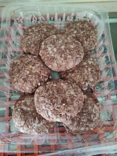 The image shows a plastic container holding eight raw, uncooked, circular patties of ground meat. The container is transparent and has condensation droplets on its surface. The patties are arranged closely together, filling the container. The meat appears to be a mix of red and white, suggesting a combination of lean and fatty ground meat. The background is indistinct but appears to be a surface of some kind. The photograph was likely taken indoors. There is no evidence of a location in Musachevo, Bulgaria.