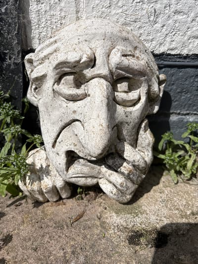 The media file captures a weathered, light-colored stone or concrete sculptural ornament in Longden, United Kingdom. The main subject is an intricately carved object, possibly depicting a stylized head or an abstract decorative element, featuring deep indentations and a rough, aged texture with visible dirt and grime from outdoor exposure.
The ornament rests outdoors against a coarse, white-painted wall, which appears to be brick or render, on the left side of the frame. Beneath the sculpture, there is a dark, rough surface. To the right, the ground consists of textured, light grey-brown concrete or paving stones. Small clusters of green plants are growing around and near the ornament, indicating a natural or slightly overgrown outdoor setting.
The scene is illuminated by bright sunlight, which casts strong, well-defined shadows from the ornament onto both the wall and the ground, suggesting a clear, sunny day. There is no discernible activity or event taking place, as the ornament is static. No visible text is present in the image.