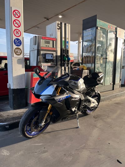 A black and silver Yamaha R1 motorcycle is parked in front of a gas station pump, pump number 5,  at a gas station located in Jaljulia, Israel at 32.146 latitude and 34.952 longitude. The motorcycle is on its kickstand. The pump is covered in a red, white, and black plastic casing.  There are various signs on the white pillar to the left of the motorcycle. There is a red car partially visible to the left of the image.  There is a glass-enclosed cabinet to the right of the motorcycle, filled with various bottles and items. The ground is asphalt. There is a small, white, faded mark on the ground in front of the motorcycle.  The sun is shining brightly and casting shadows.