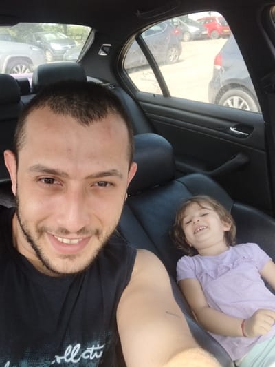 A man and a young girl are in the back seat of a car. The man is smiling at the camera while the girl is lying down and also smiling. The car is parked in a parking lot with other vehicles visible through the window. The location is inferred to be Stafford, United Kingdom.