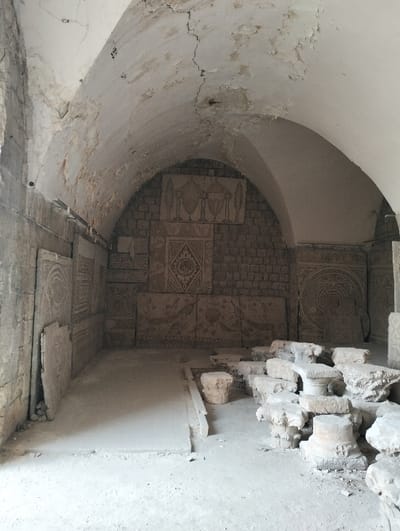 A weathered stone chamber features an arched ceiling with cracked paint and exposed brick. Carved stone panels adorn the walls, displaying intricate geometric and figurative designs. Scattered fragments of stone columns lie on the floor. The room's interior is bathed in diffuse light, highlighting the aging textures of the stone and the overall decay.