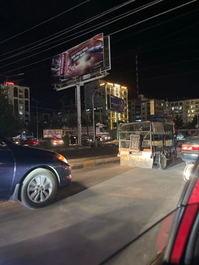 The image depicts a night scene in Kabul, Afghanistan, likely on a busy street. The foreground is dominated by the side and wheel of a dark blue car, suggesting a first-person perspective from within a vehicle. In the midground, a peculiar caged vehicle, possibly a modified truck or cart, is visible, its cage filled with what appear to be animals, or possibly goods covered by tarps. The vehicle's rough exterior and mismatched parts indicate it is a utilitarian mode of transport.

Further back, traffic is evident, including red and white cars and a larger truck, suggesting a traffic jam or slow-moving traffic. Buildings line the street, some illuminated with multiple stories and windows, displaying modern architecture. A prominent billboard is mounted on a tall structure, advertising a product with images and text. Overhead, power lines crisscross the sky, a common feature in urban environments. The overall impression is of a bustling, somewhat chaotic urban environment at night, characteristic of a developing city. The presence of the caged vehicle is a unique element that stands out.