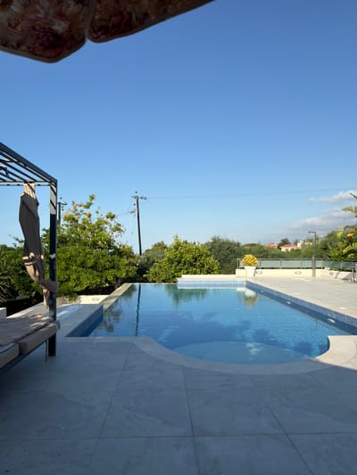 A bright, sunny day is captured in this image of a resort-style swimming pool area. The pool itself is rectangular with a curved section, filled with clear blue water that reflects the sky and surrounding trees. The pool is surrounded by a light-colored tiled deck. To the left, a portion of a lounge chair with a beige cushion is visible under a dark metal pergola. A folded beige umbrella hangs from the pergola. Lush green trees and some flowering bushes are in the background, along with power lines and poles. In the distance, houses with red roofs are nestled amongst the trees. The sky is a clear, bright blue, with a few wispy clouds towards the right side of the frame. The overall impression is one of a well-maintained, tranquil outdoor living space, likely in a warm climate such as Kyrenia, Northern Cyprus.