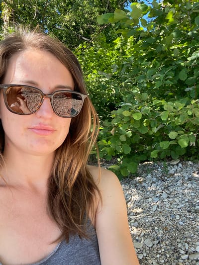 A young woman with long brown hair and sunglasses is taking a selfie outdoors. She is wearing a grey tank top. The background consists of lush green trees and a rocky shore. The sun is shining brightly, casting shadows and highlighting the textures of the environment. The reflection in her sunglasses shows a hand and the rocky ground. The scene appears to be a natural setting, likely a park or a lakeside area in Cortaillod, Switzerland, given the lush vegetation and the presence of a body of water suggested by the rocky shore.