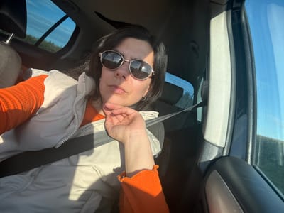 A person with dark hair, wearing sunglasses and a seatbelt, is in a car. They are wearing an orange top and a light-colored vest. The location is Breckland District, United Kingdom, suggested by the reflection of UK buildings on the sunglasses.