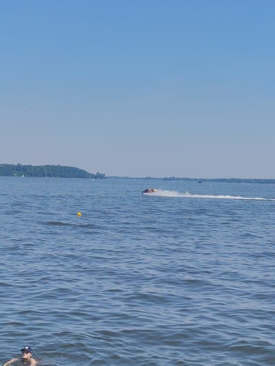 The image captures a bright, clear day at a body of water, likely Lake Zegrzyńskie near Nieporęt, Poland. The sky is a solid, pale blue, and the water is a deep blue with gentle ripples. In the foreground, a person is partially submerged in the water, wearing a blue cap. Further out on the water, a yellow buoy bobs. In the mid-ground, a jet ski is speeding across the water, leaving a white wake. In the background, a tree-covered shoreline stretches across the horizon, with several sailboats dotted on the water and in the distance.