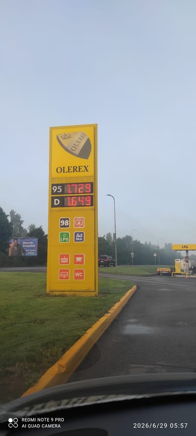This is a photograph taken from inside a vehicle looking towards a gas station in Maardu, Estonia. The primary focus is a tall yellow price sign for "OLEREX" gas station. The sign displays fuel prices: 95 is 1.729, and D is 1.649. Below these, there are icons representing 98 octane fuel, 24-hour service, LPG, AdBlue, a shopping basket, a coffee cup, a caravan, and a WC. To the left of the sign, a billboard advertises "Maardu linnapeaiv" with an image of a man. In the background, trees line the road, and a black SUV is visible driving away from the gas station. Further in the distance, a small trailer is parked near an LPG gas pump. The sky is overcast, and the ground appears wet, suggesting recent rain. The image also contains a timestamp "2026/6/29 05:57" and "REDMI NOTE 9 PRO AI QUAD CAMERA" at the bottom left corner.