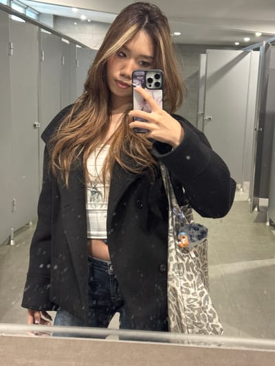 A young woman is taking a mirror selfie in what appears to be a public restroom. The restroom is illuminated by overhead lights. The stalls are gray and metallic. She is holding up a smartphone, an iPhone, in her right hand. She is wearing a black coat over a white top. She is also wearing blue jeans. She has long, light-brown hair. The scene is not in Guazhou County, China.
