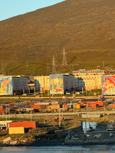 The image captures a view of the city of Pevek, Russia, characterized by its distinctive Arctic landscape and urban development. In the foreground, the water's edge is visible, with debris and concrete structures suggesting a port or industrial area. Several shipping containers, some brightly colored in red and yellow, are stacked and arranged on the shore, indicating ongoing logistics and trade. Further back, a series of low-rise buildings, possibly residential or commercial, are visible, some featuring murals of animal imagery, such as a bear or deer. These structures are interspersed with industrial elements like cranes and power lines. The backdrop is dominated by a large, barren hill or mountain, its slopes covered in sparse vegetation and casting long shadows in the warm, low sunlight. The sky is clear and blue, typical of a sunny day. The overall impression is one of a remote, working town situated in a stark natural environment, with a blend of functional infrastructure and decorative elements.