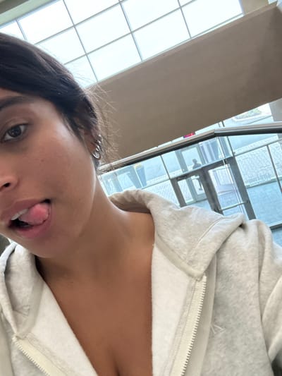 The image depicts a close-up selfie of a female individual, foregrounded, with her dark hair pulled back. She has tan skin, dark eyes, and multiple silver hoop earrings in her visible left ear. Her mouth is slightly open, and her tongue is partially extended. She is wearing a light grey, full-zip hooded sweatshirt, which is unzipped to reveal her chest.

The background shows a contemporary indoor environment. Overhead, a large grid-patterned skylight ceiling allows natural light. Below the ceiling, a wide, light-colored (possibly beige or off-white) architectural beam or wall runs horizontally. Further back, large glass windows or panels provide a view to an exterior paved area and reflect interior elements. A faint silhouette of an individual, possibly in a uniform, is visible through the glass near an entry/exit point, above which a red "EXIT" sign is partially discernible. The overall architectural style, including the expansive glass and public area appearance, strongly indicates the Penn Medicine Princeton Medical Center, located in Plainsboro Township, New Jersey, United States.