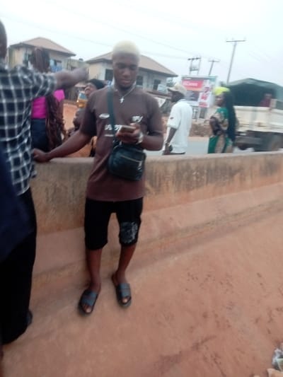 The image captures an outdoor scene, likely along a street or roadside in a residential or semi-urban area, featuring several individuals.
In the center foreground, a young man with dark skin stands prominently. He has striking light, possibly bleached or dyed, blonde hair cut short. He is wearing a dark brown short-sleeved t-shirt with some indistinct light-colored graphics or text on the front. Below the waist, he wears dark shorts, which appear to be black, with a patterned or graphic patch on the lower right leg. On his feet, he has dark, open-toed slide sandals. A dark messenger-style bag or satchel is slung across his chest, resting on his right hip. Around his neck, he wears a silver-colored chain with a small cross pendant. He is holding a smartphone in both hands at chest level, appearing to be looking intently at its screen.
To the left of the central figure, a person wearing a dark and white checkered long-sleeved shirt is partially visible, with their left arm extended outwards. Further back and slightly to the left, another person with long dark hair, wearing a bright pink top, is partially seen behind the checkered-shirt individual.
In the mid-ground, to the right of the central figure, a man in a white short-sleeved collared shirt and a light-colored baseball cap is visible, facing away from the camera. Further to the right, a person, possibly a woman, is seen in profile wearing a long, flowing dress with a vibrant green and possibly yellow/brown pattern. This person also appears to have bright, possibly blonde or dyed, hair. Behind these figures, a white truck is partially visible on what seems to be a paved road.
The central figure and the person in the checkered shirt are positioned on a wide, reddish-brown concrete or dirt-covered raised barrier or ledge that runs horizontally across the lower part of the image. The surface of this barrier shows some dirt, minor stains, and texture variations.
In the background, beyond the road, there are buildings with reddish-brown roofs, indicating a built-up area. Overhead, power lines are visible against a light, overcast sky, suggesting either an overcast day or late afternoon/early evening lighting conditions. A billboard or large sign with some faint, unreadable text and graphics can be seen in the far right background, above the road. The overall impression is one of a casual, everyday street scene with people going about their activities.