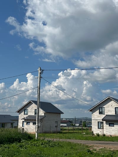 The image captures a scene in a semi-rural area, possibly a developing residential zone, with two unfinished houses constructed from light-colored blocks and dark roofs. The houses are situated on a slight incline, with a dirt path in the foreground leading towards them. Lush green grass and some scattered vegetation are visible in the foreground and around the houses. A concrete utility pole with overhead wires stands prominently in the center-left of the frame, interrupting the view of the sky. The sky itself is a dynamic mix of bright blue and large, billowy white and grey clouds, suggesting a day with changeable weather. In the background, a distant, hazy urban or suburban landscape with more buildings and greenery is visible. There are no people or animals present in the image. The overall impression is one of a construction site or a newly developing neighborhood under a dramatic sky.