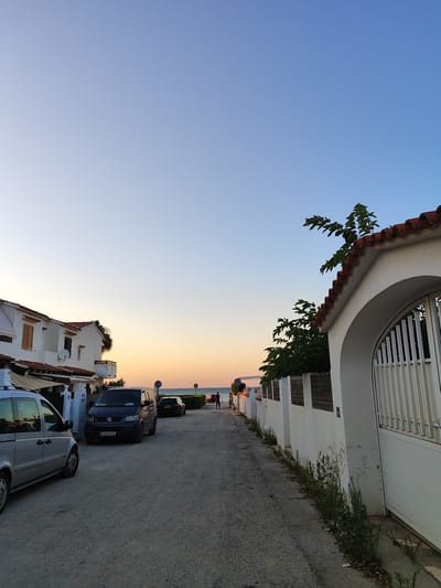 A street scene in Els Poblets, Spain, at dusk. The sky transitions from a clear, pale blue at the top to warm orange and yellow hues near the horizon, indicating the setting sun. On the left, a row of white buildings with terracotta tiled roofs line the street, with a silver minivan parked in the foreground. To the right of the street, a white wall with a large arched gateway and a white metal gate stands, with green foliage growing along its base. Further down the street, a dark blue van is parked, followed by a black car and a distant view of the sea and beach. Two figures are walking away from the camera down the street towards the beach. The street is paved with asphalt.