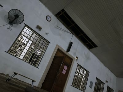 The image depicts the interior of a building in Abeokuta, Nigeria. The walls are white, and there are multiple windows with decorative metal grilles. A dark wooden door stands ajar, adorned with two pink papers. A large, industrial fan is mounted on the wall near a window. An analog clock hangs higher on the wall above the window. A folding table sits in front of the window. A speaker is vertically mounted on the wall next to the door. The ceiling is comprised of gray panels with a long rectangular opening.