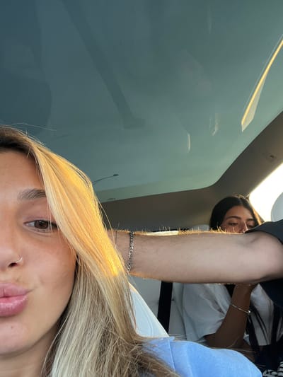 The image depicts the interior of a vehicle, located in Kochav Yair, Israel. In the foreground, the left side of a light-skinned female's face is visible, characterized by long blonde hair, a brown eye, and a small silver ring piercing on her left nostril. Her lips are pursed. Golden sunlight illuminates the left side of her hair. A light blue garment is partially visible on her lower left. From the mid-right, an arm with visible hair and a silver Figaro-style chain bracelet extends horizontally, partially obscuring the background. Seated in the rear of the vehicle, a second light-skinned female is visible with dark hair, wearing a white t-shirt and a thin silver bracelet on her right wrist; her head is oriented downwards. The vehicle interior features a panoramic glass roof, reflecting the sky and internal structural components.