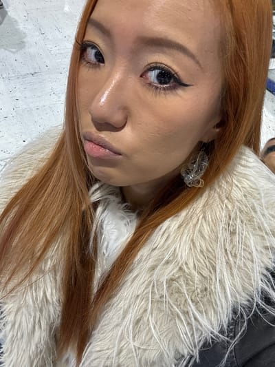 A striking portrait of a woman with light brown hair, adorned with a fluffy white faux fur collar. She gazes upwards with glossy, lined eyes and a subtle, pink lip. Delicate, transparent earrings accentuate her features. The lighting highlights the contours of her face, giving it a radiant glow. The image has a soft, slightly tilted perspective.