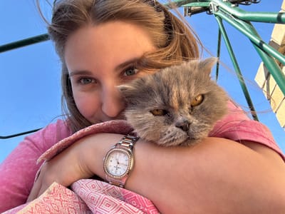 A young woman is holding a gray cat, viewed from a low angle against a bright blue sky. The woman has fair skin, light brown hair, and is wearing a pink t-shirt and a pink and white patterned shawl. She is wearing a watch on her left wrist. The cat is gray with amber eyes and its face is close to the woman's neck. Green metal poles are visible in the lower left corner. The location is Druzhino, Russia.