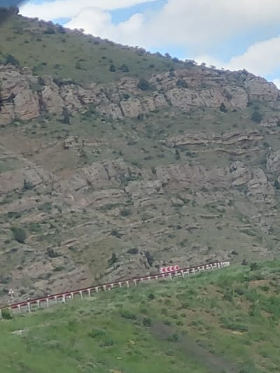 This image captures a rugged mountainous landscape, likely situated in Tigranashen, Armenia. The scene is dominated by a steep, rocky mountainside covered with patches of green vegetation and sparse bushes. The geological formations consist of large, greyish-brown rocks with visible stratification and cracks.

In the upper and lower left portions of the tilted frame, segments of a bright blue sky with white, fluffy clouds are visible, indicating a daytime setting, possibly under partly cloudy conditions. The lighting suggests it is bright, likely during late morning or afternoon.

A prominent feature in the lower right half of the image is a road barrier, stretching diagonally across the frame. This barrier is characterized by a repeating pattern of red and white chevron markings, typically used to indicate a curve, hazard, or edge of a road. Adjacent to the barrier, the ground is covered with lush green grass and some darker patches of shrubbery.

The image is captured with a noticeable tilt, presenting the mountain face and the barrier at an angle rather than horizontally or vertically. No people or specific activities are discernable within the frame, focusing solely on the natural and man-made elements of the landscape. The scene conveys a sense of a remote, natural environment traversed by a road, common in mountainous regions.