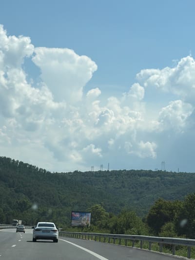 This is a landscape photograph taken from a moving vehicle on a highway. The foreground shows a multi-lane asphalt road with a white car in the right lane, a gray car in the left lane, and a truck further ahead in the left lane. A metal guardrail lines the right side of the road. The midground features a dense, green, tree-covered hillside. In the background, a bright blue sky is filled with large, white, puffy clouds. Power lines and pylons are visible on the crest of the hill. A billboard with images and text is mounted on the hillside to the right of the road. The scene appears to be in a rural or semi-rural area, possibly near Churek, Bulgaria, given the natural landscape and the presence of power infrastructure. The lighting suggests it is daytime.