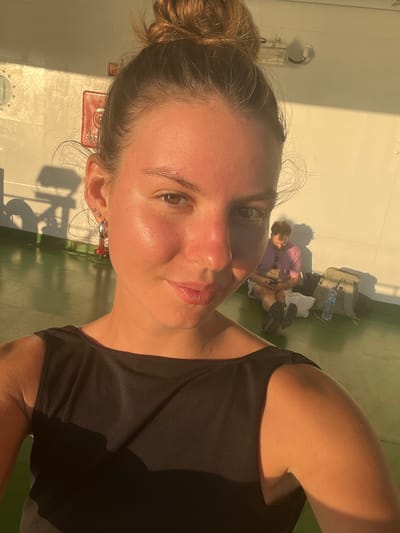 The image shows a young woman with her hair in a bun, looking directly at the camera. She is wearing a black sleeveless top and large hoop earrings. Her skin is illuminated by warm, golden light. In the background, out of focus, a man is sitting on the floor, looking at his phone. He is wearing a purple shirt and shorts. There is a backpack and a water bottle next to him. The environment appears to be an outdoor deck area with a white wall and a green floor. The scene is bathed in the warm glow of what appears to be sunset or sunrise. The image is a selfie taken by the woman in the foreground. The location is not specified as Batas Desa Skotong Timur, Indonesia.