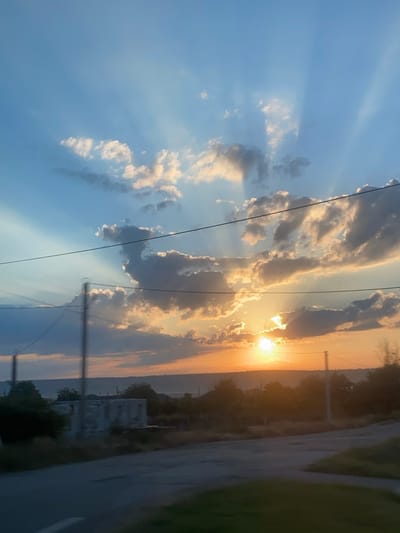 The image depicts a sunset over a rural landscape, possibly in or near the city of Svetlîi, Moldova. The sky is filled with a dramatic display of light and clouds, with the sun partially obscured by clouds, casting rays of light downwards. The foreground shows a road and some vegetation. In the middle ground, there are trees and an unfinished or dilapidated building, suggesting an area under development or in disrepair. The overall scene conveys a sense of dusk in a somewhat undeveloped rural setting.