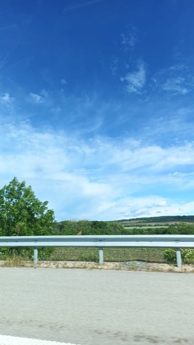 A daytime, outdoor scene shows a road with a gray guardrail running along the edge. Beyond the guardrail, there is a green field with low-lying vegetation and trees. Further in the distance, there is a hilly landscape with patches of trees and fields. The sky is blue with scattered white clouds. No people or other discernible subjects are present. The location details are Yazdach, Bulgaria.