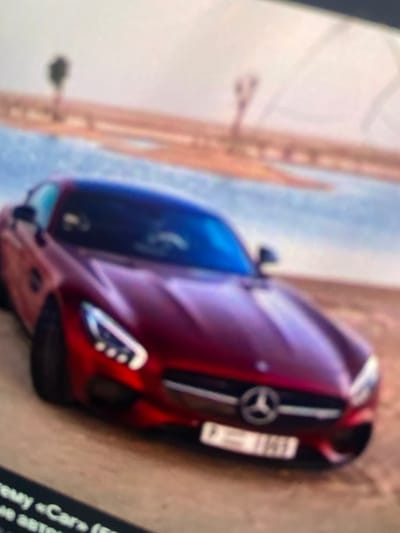 A blurred, red Mercedes-Benz car is parked on a sandy surface, possibly a desert road. The car has a distinctive grille with the Mercedes-Benz logo and a license plate that appears to be from the United Arab Emirates. In the background, there is a body of water and a sandy landscape with sparse vegetation, suggesting an arid or desert environment. The sky is overcast. The image quality is low, with significant blur. No people are visible in the image. The lower left corner shows a fragment of text, which is illegible due to the blur. The location in Paczkowo, Poland is not depicted in this image.