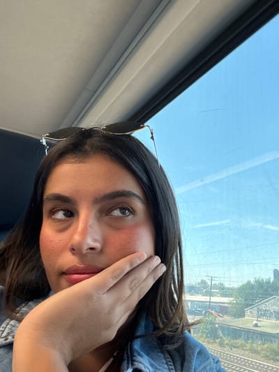 A young adult female, with dark hair and light brown skin, is captured from the chest up inside a train car. She looks upwards and to the right, her right hand resting against her right cheek. Dark-lensed sunglasses are perched on top of her head. Through the train window, a clear blue sky is visible. Below the sky, an industrial landscape unfolds, featuring multiple parallel railroad tracks. Beyond the tracks, green foliage, utility poles with power lines, and several industrial buildings are present. A freight train with a partially visible "MAERSK" logo on a red cargo container is discernible in the mid-ground. One industrial structure displays visible graffiti. This scene is located within the city of Newark, United States, likely along an active rail corridor passing through its industrial and port areas.