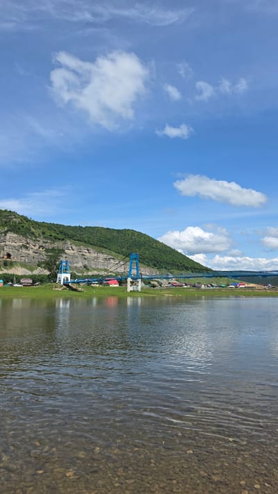 The image captures a scenic view of a body of water with a mountainous, wooded landscape on the left, and a clear blue sky with scattered white clouds above. A suspension bridge, painted in a vibrant blue, spans across the scene, with its towers anchored to the rocky mountainside. Along the base of the mountain, a line of colorful buildings, possibly homes or small establishments, are visible. The water appears to be shallow near the shore, revealing a rocky bottom, and then deepens to a reflective surface that mirrors the sky and surrounding elements. The sun appears to be high in the sky, suggesting it is daytime, and the weather seems pleasant. The setting, identified as Tash-Asty, Russia, evokes a sense of natural beauty and perhaps a touch of rural or recreational development. No people are distinctly visible, but the presence of buildings suggests human habitation or activity. The overall mood is tranquil and picturesque.