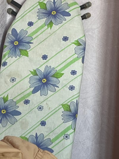 The image shows a close-up of an ironing board cover with a pattern of blue flowers and green stripes. The ironing board is leaning against a light purple textured fabric, possibly a curtain or wall covering. Two dark rods with grey caps are visible at the top edge of the ironing board. A portion of a person's arm or shoulder, covered in beige fabric, is in the lower left corner of the frame. There are also some brown stains on the ironing board cover. The location in the city of Gari, Russia is not discernible from the visual information provided in the image.